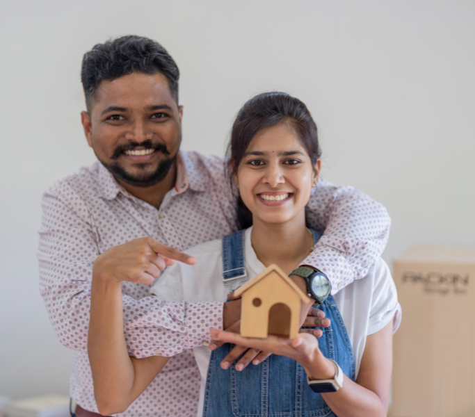 Young smiling couple holding and pointing to a small wooden house